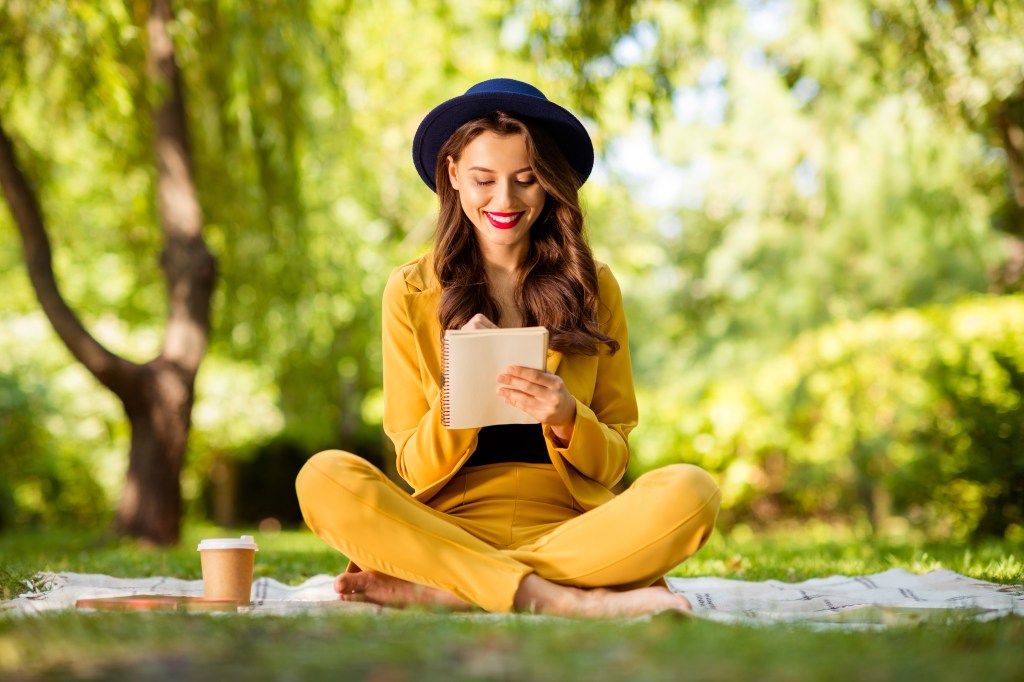 Cheerful woman with wavy hair sitting in a lotus position on a blanket outside, taking notes